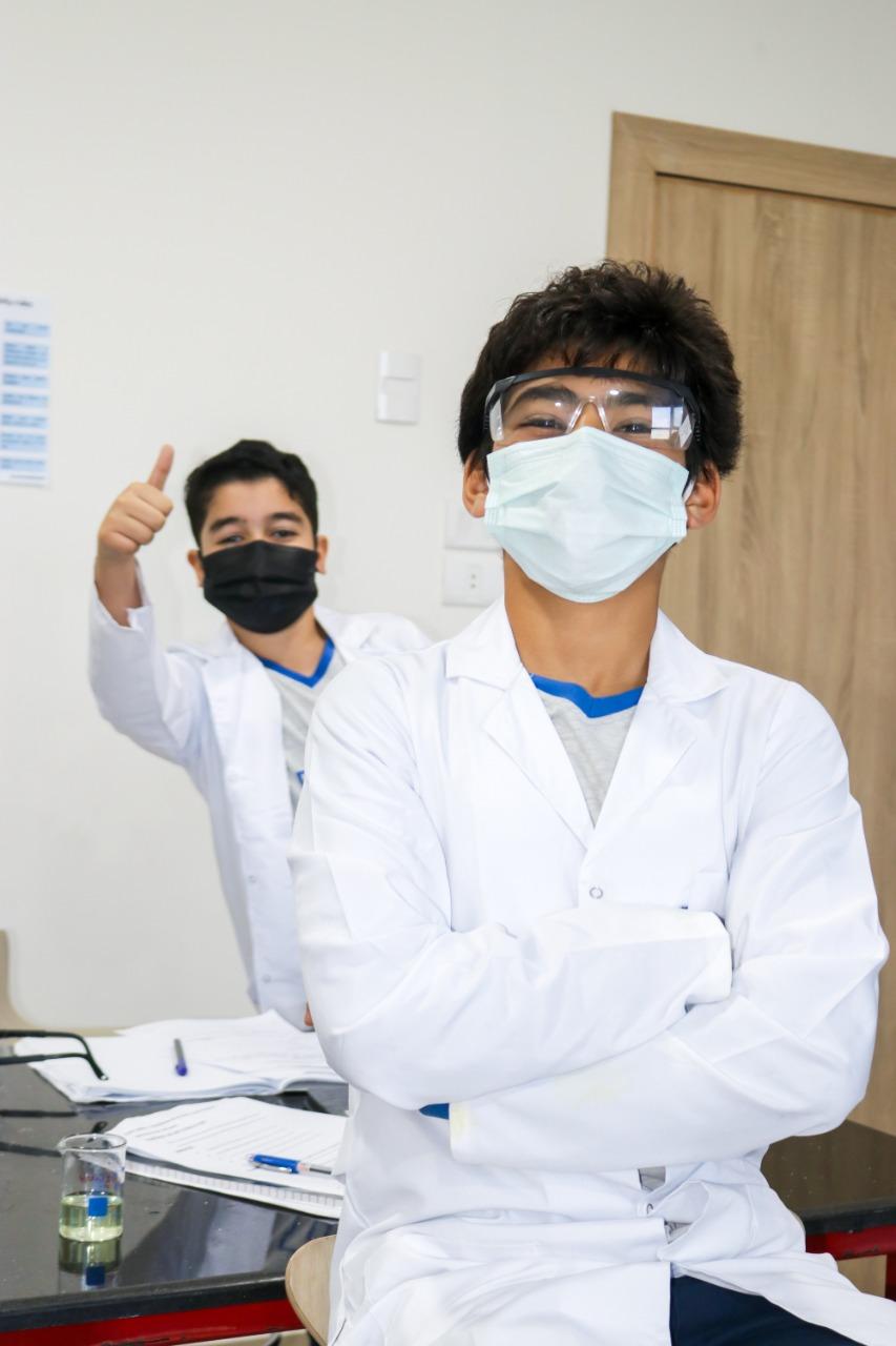 Experience hands-on laboratory work at IVY STEM International School. Two individuals in lab coats, safety goggles, and face masks are seen confidently conducting experiments. A blue liquid-filled beaker and educational materials are visible on the table.